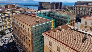 A cityscape with a prominent castle-like structure in the background, modern buildings with visible construction scaffolding, and a clear view of the sea. - Art Flat Municipio (Naples)