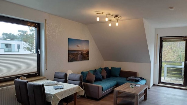 A modern living room with a sofa, chairs, a coffee table, and a dining table. There is a painting on the wall and a window with a view of a building outside. - Vacation apartment with dog in Cuxhaven Sahlenburg Apartment 3 (Cuxhaven)