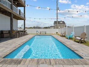 A rectangular swimming pool with a wooden deck surrounding it, a wooden deck with a railing, and string lights above.