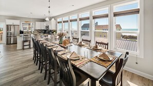 Een ruime eethoek met een lange tafel voor zes personen, een keuken op de achtergrond en grote ramen met uitzicht op het strand.
