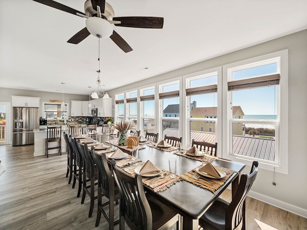 A spacious dining area with a long table set for six, a kitchen in the background, and large windows offering a view of the beach.