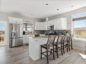 A modern kitchen with white cabinets, stainless steel appliances, a central island with bar stools, and a view of the city through large windows. - Pasture to Paradise #126 - Private Pool, Ocean Views, Dog Friendly, Elevator! (North Topsail Beach)