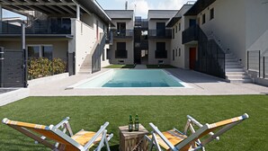 Un immeuble résidentiel moderne doté d’une piscine centrale, de deux chaises longues et d’une petite table en bois sur laquelle sont posées des bouteilles.