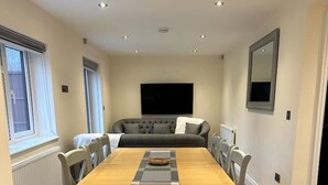 A dining area with a wooden table, grey upholstered chairs, a grey sofa, and a flat-screen TV mounted on the wall. - The Grey Retreat (Shepperton)