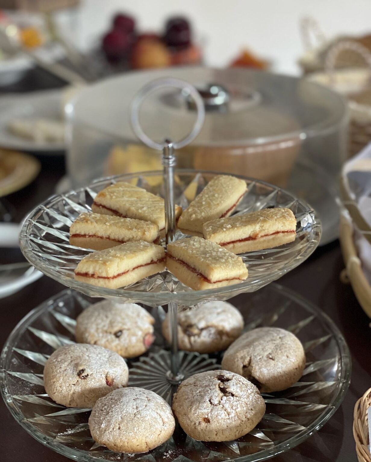 A multi-tiered glass serving tray with pastries and a glass container in the background.
