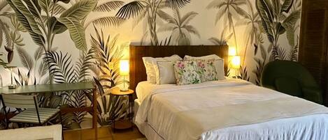 A bedroom with a bed, two bedside tables with lamps, a chair, a table, and a wall with a tropical plant mural.