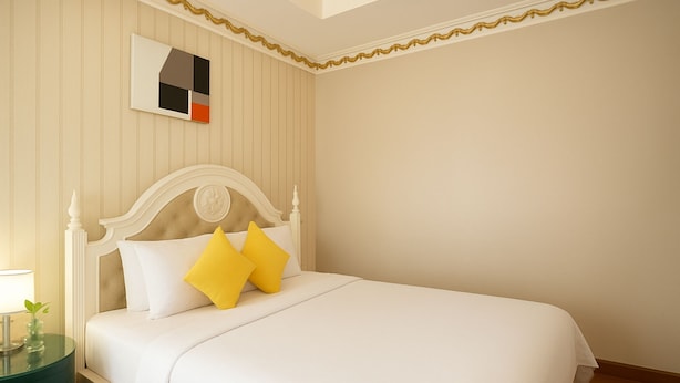 A neatly made bed with white bedding and two yellow pillows, a bedside table with a lamp, and a framed abstract artwork on the wall. - HANZ 4013 Hotel Saigon (Ho Chi Minh City)