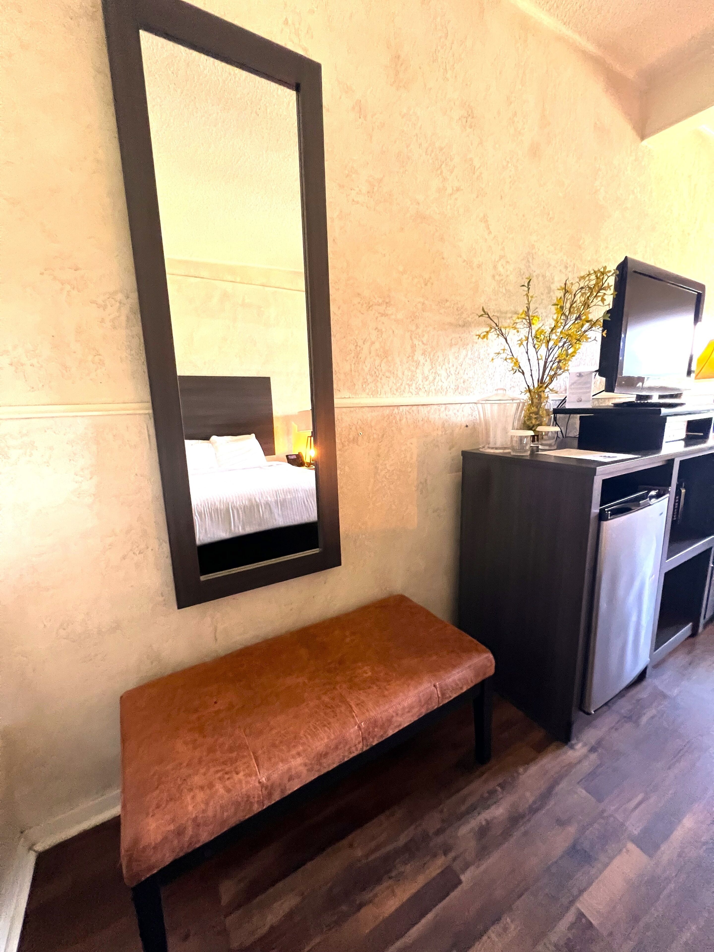 Hotellihuone, jossa on suuri peili, puinen penkki, pieni pöytä maljakolla, televisio ja jääkaappi.
