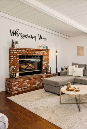 A living room with a brick fireplace, a grey sofa, a round coffee table, and a wall with the text 'Whispering Wine'.