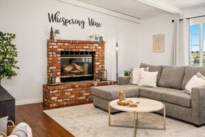 A cozy living room with a brick fireplace, a grey sofa, a round coffee table, and a large window with a view.