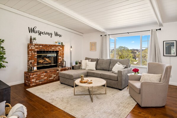 A living room with a brick fireplace, a grey sectional sofa, a round coffee table, and a large window with a view of trees.
