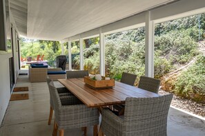 A covered patio with a wooden table and wicker chairs, overlooking a lush garden.