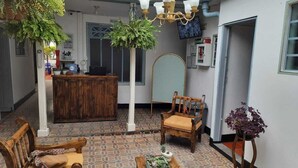 A reception area with a wooden desk, two wooden chairs, a hanging plant, a chandelier, and a tiled floor. - Hotel Posada Santa Elena (Tunja)