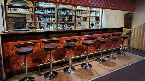 Uma área de bar com balcão estofado vermelho, várias prateleiras cheias de garrafas e seis banquetas.