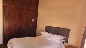 Cama bem arrumada com lençóis brancos em quarto com guarda-roupa de madeira e pequeno armário de madeira.
