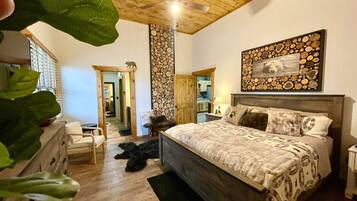 Une chambre avec un plafond en bois, un lit recouvert d’une literie à motifs, un tableau accroché au mur et un plancher en bois.