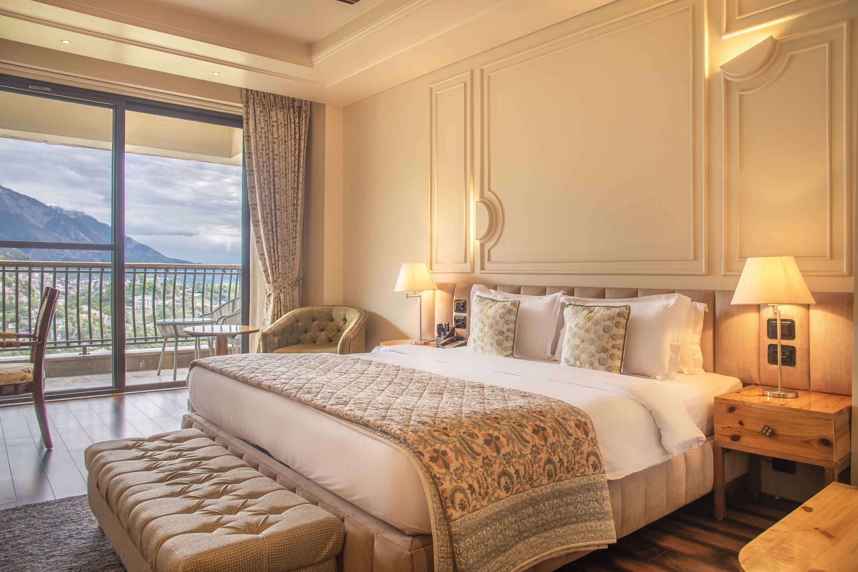A spacious bedroom with a large bed, two bedside tables with lamps, a chair, and a balcony with a view of mountains.