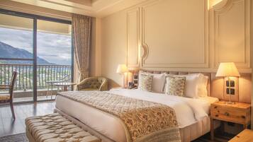 A spacious bedroom with a large bed, two bedside tables with lamps, a chair, and a balcony with a view of mountains.