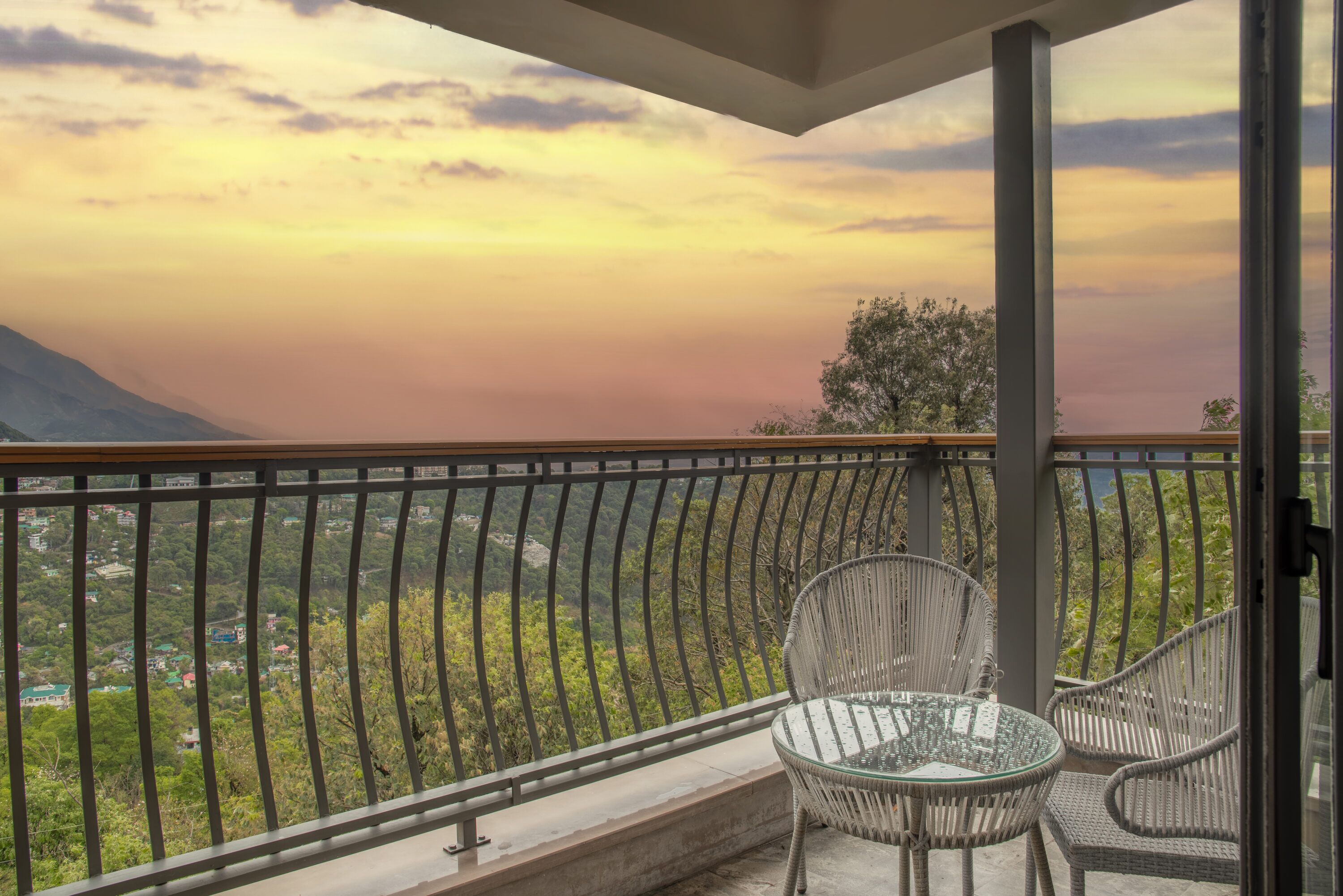 A balcony with a glass table and chairs, overlooking a mountainous landscape.