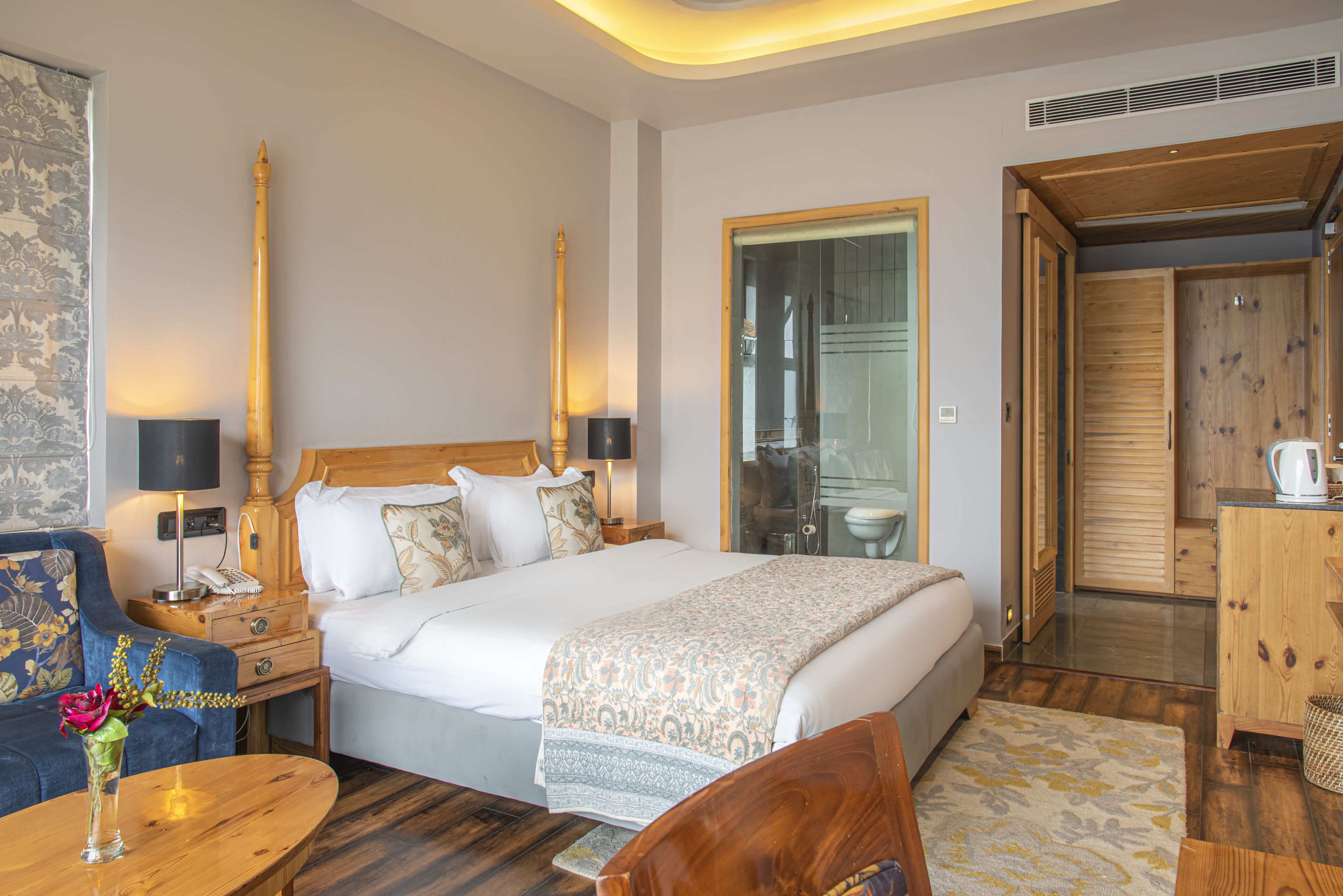 Habitación de hotel con una cama grande, cabecera de madera, mesitas de noche con lámparas, una pequeña mesa redonda con un florero, un sillón azul y un armario de madera.