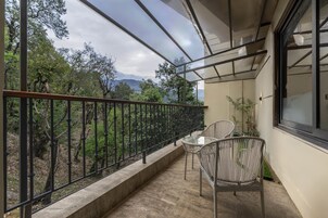 Ein Balkon mit Metallgeländer, zwei Korbstühle, ein kleiner Tisch und Blick auf Bäume und Berge.