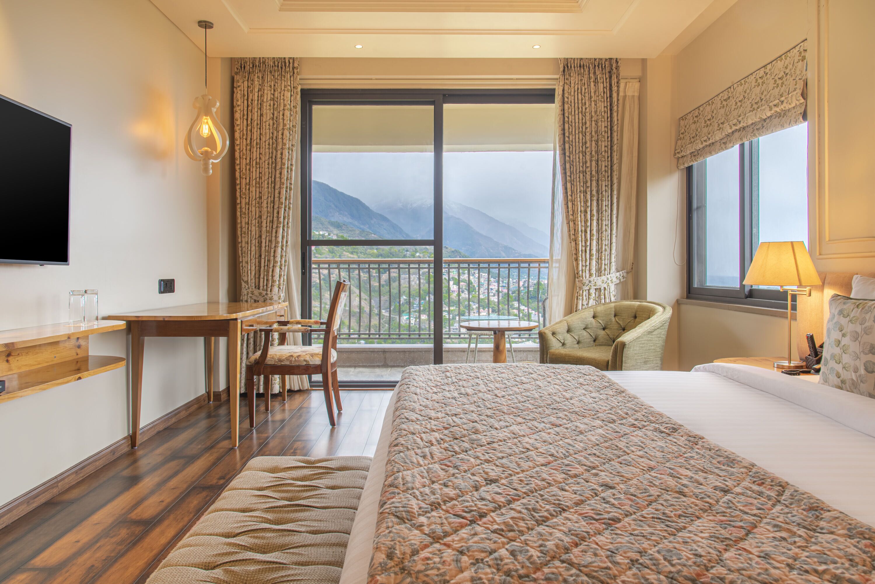A hotel room with a large bed, a desk, a chair, a TV, and a balcony with a mountain view.