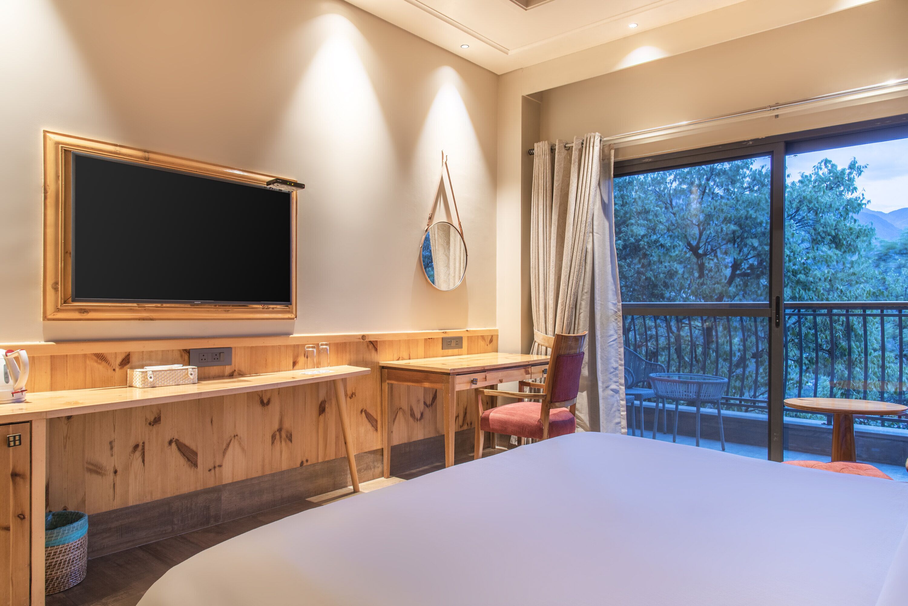 A hotel room with a large bed, a wooden desk, a flat-screen TV, a chair, a small table, and a balcony with a view of trees and mountains.