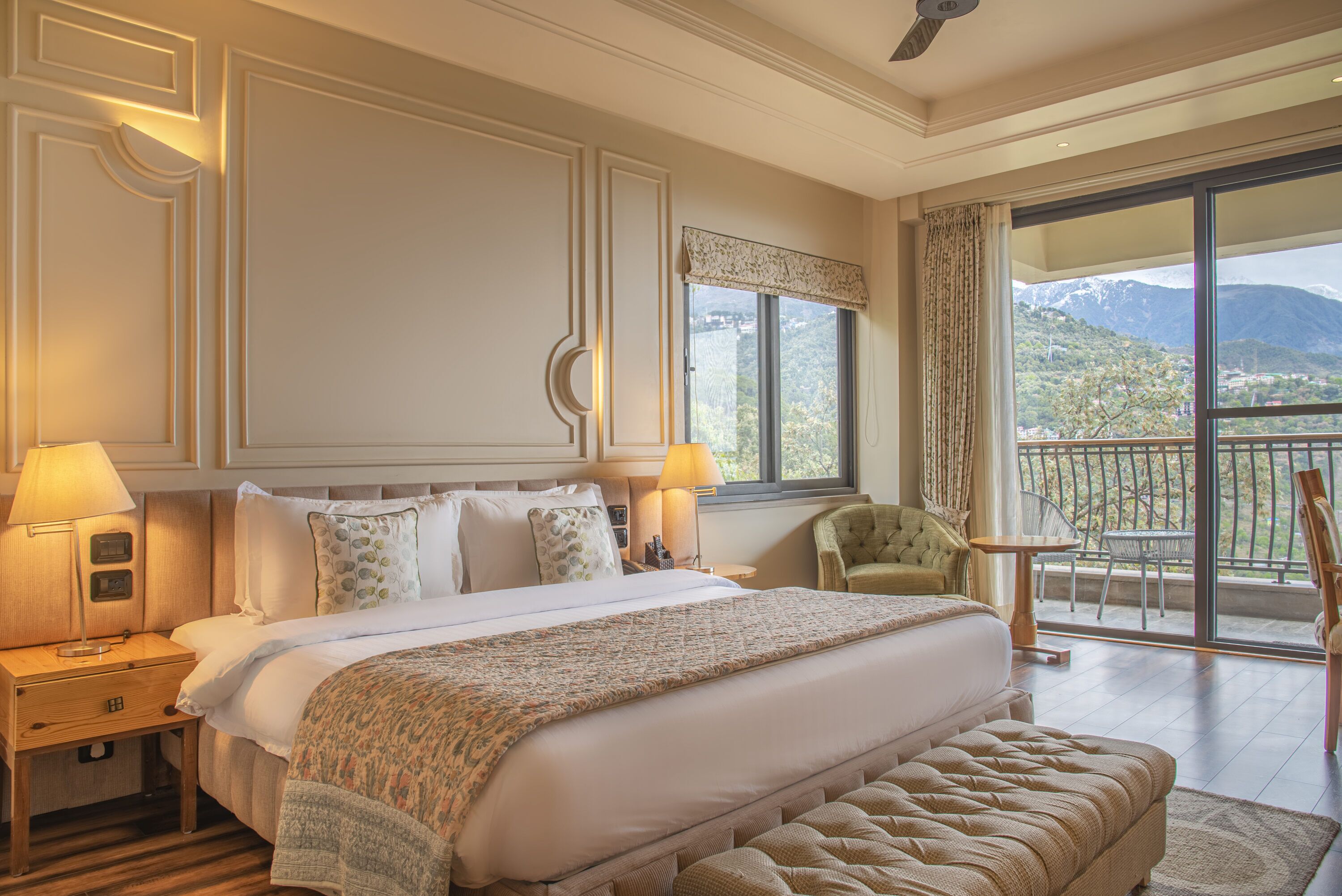 Une chambre d’hôtel avec un grand lit, deux lampes de chevet, un fauteuil vert, une petite table et un balcon offrant une vue sur les montagnes.