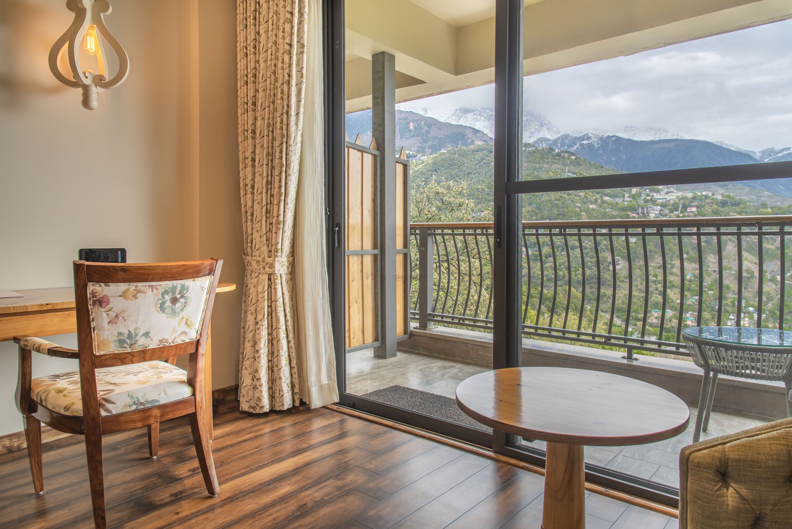 A room with a wooden chair, a round table, and a view of mountains through a balcony.