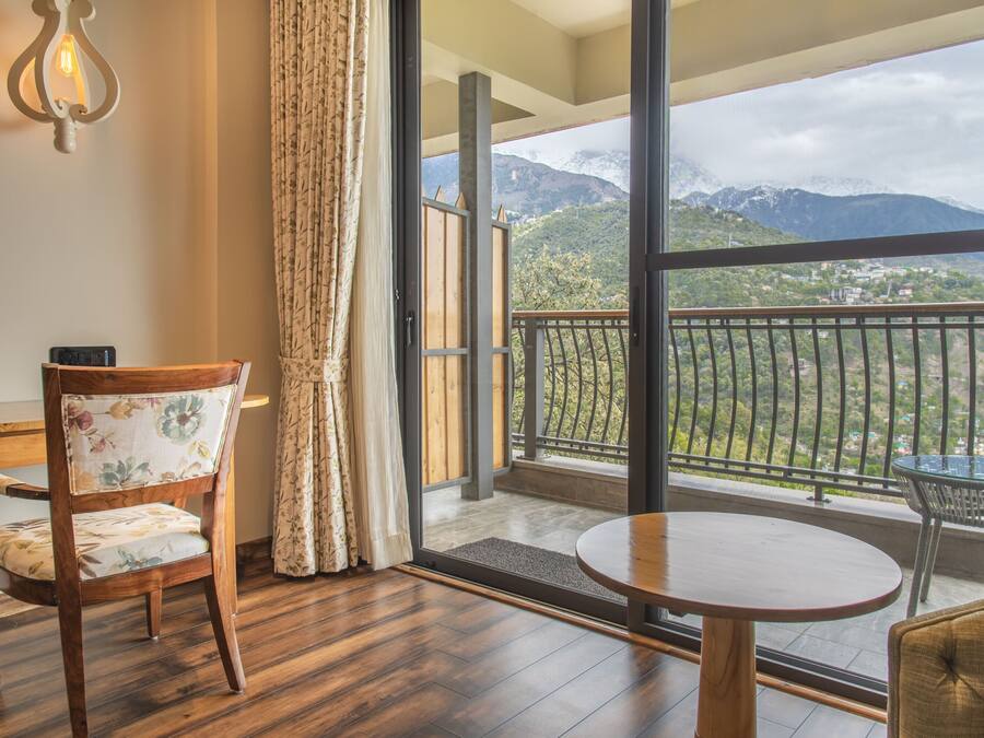 A room with a wooden chair, a round table, and a view of mountains through a balcony.