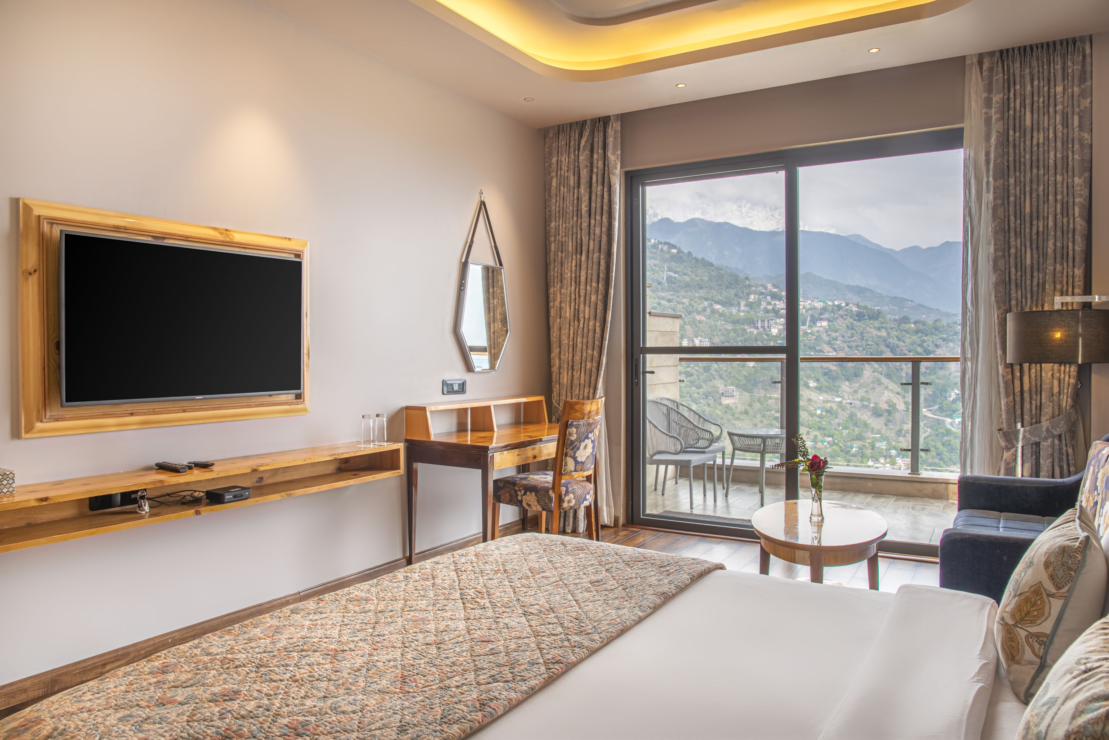 A modern hotel room with a large bed, a TV mounted on the wall, a desk with a chair, a small table with a vase, and a balcony with a view of mountains.