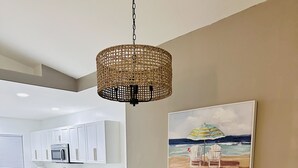 A dining area with a wooden table and chairs, a woven pendant light, and a beach-themed painting on the wall.