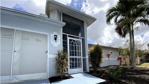 A modern two-story house with a garage, a front door with glass panels, and a small garden with palm trees and shrubs.