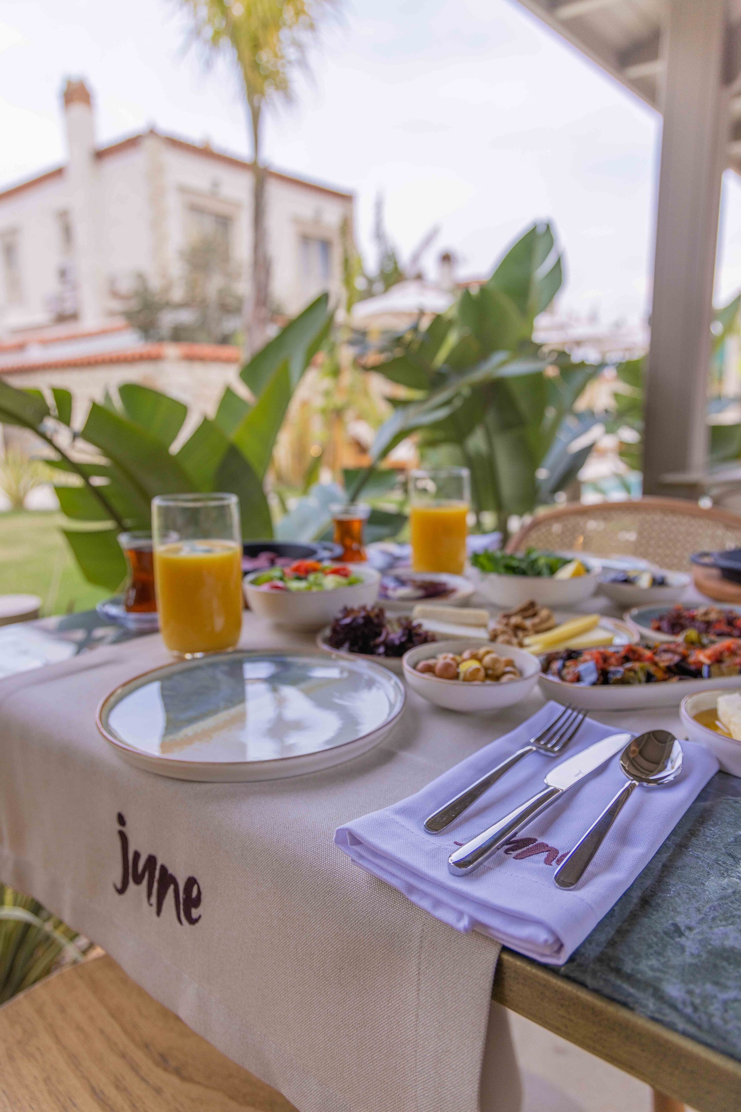 Une table de petit-déjeuner installée à l’extérieur, avec différents plats, des verres de jus d’orange et des couverts soigneusement disposés.