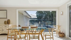 Un coin repas avec une table en verre et des chaises en bois, un meuble blanc agrémenté d’objets décoratifs, et une vue sur une piscine à travers de grandes portes vitrées.