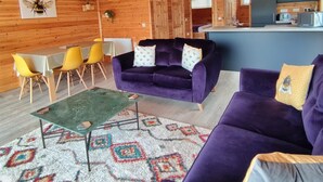 A spacious wooden cabin with a dining area, two purple sofas, a kitchen, and a ceiling with hanging plants. - Nature's Nook - Norfolk Broads (Brundall)