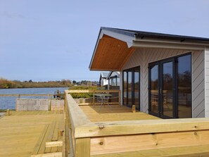 A modern house with a deck overlooking a body of water, featuring large sliding glass doors and a wooden deck.