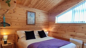 A wooden cabin bedroom with a bed, bedside lamp, and a window with blinds. - Nature's Nook - Norfolk Broads (Brundall)