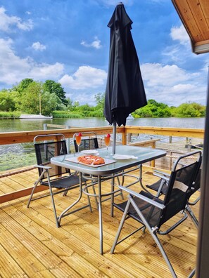 A wooden deck with a table and chairs, a black umbrella, and a view of a river with boats.