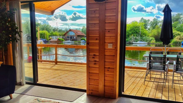 A wooden interior with a large window overlooking a deck and a lake. - Nature's Nook - Norfolk Broads (Brundall)