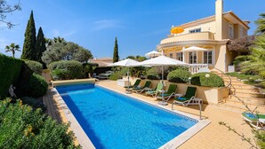 Ein Swimmingpool mit Liegestühlen und Sonnenschirmen, umgeben von gepflegter Bepflanzung und einem großen Gebäude mit einem Ziegeldach.