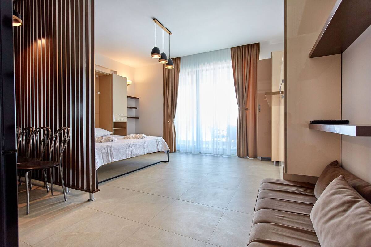 Una habitación de hotel moderna con una cama, una zona de estar con sofá, una mesa de comedor con sillas y un gran ventanal con cortinas.