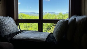 Habitación con un ventanal grande que ofrece vistas a árboles verdes y montañas. Cuenta con una cama con ropa blanca y un almohadón mullido.