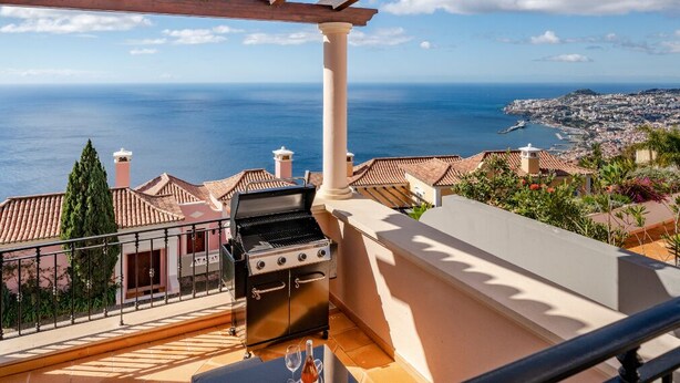 A balcony with a grill, a table with a glass, and a view of the sea and city. - Palheiro Village villa, priv pool | Palheiro Ocean (São Gonçalo)