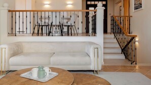 A living room with a sofa, a coffee table with mugs, a staircase with a railing, and a dining area in the background. - Palheiro Village villa, priv pool | Palheiro Ocean (São Gonçalo)