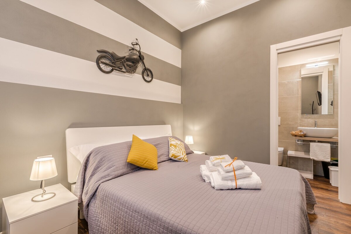 Quarto com cama, mesa de cabeceira com abajur, motocicleta na parede e banheiro com pia e espelho.