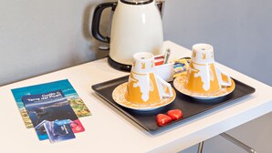Una mesa con una cafetera, dos tazas decoradas, un reposabajares, folletos y un plato pequeño con dos objetos.