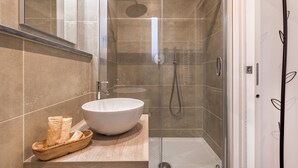 Un baño moderno con un lavabo blanco de diseño, un cuenco de madera con toallas y una zona de ducha con mampara de vidrio.