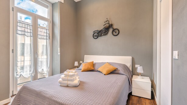 Un dormitorio con cama, decoración de motocicleta en la pared, ventana con vista a edificios y puerta con cortina.