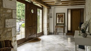 Una habitación amplia con una gran puerta de madera, una pared de piedra, piso de baldosas, un banco de madera y una mesa blanca con un florero.